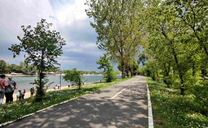 Biciklistička staza na Adi Ciganliji, okružena zelenilom, pored jezera sa bistrom vodom.