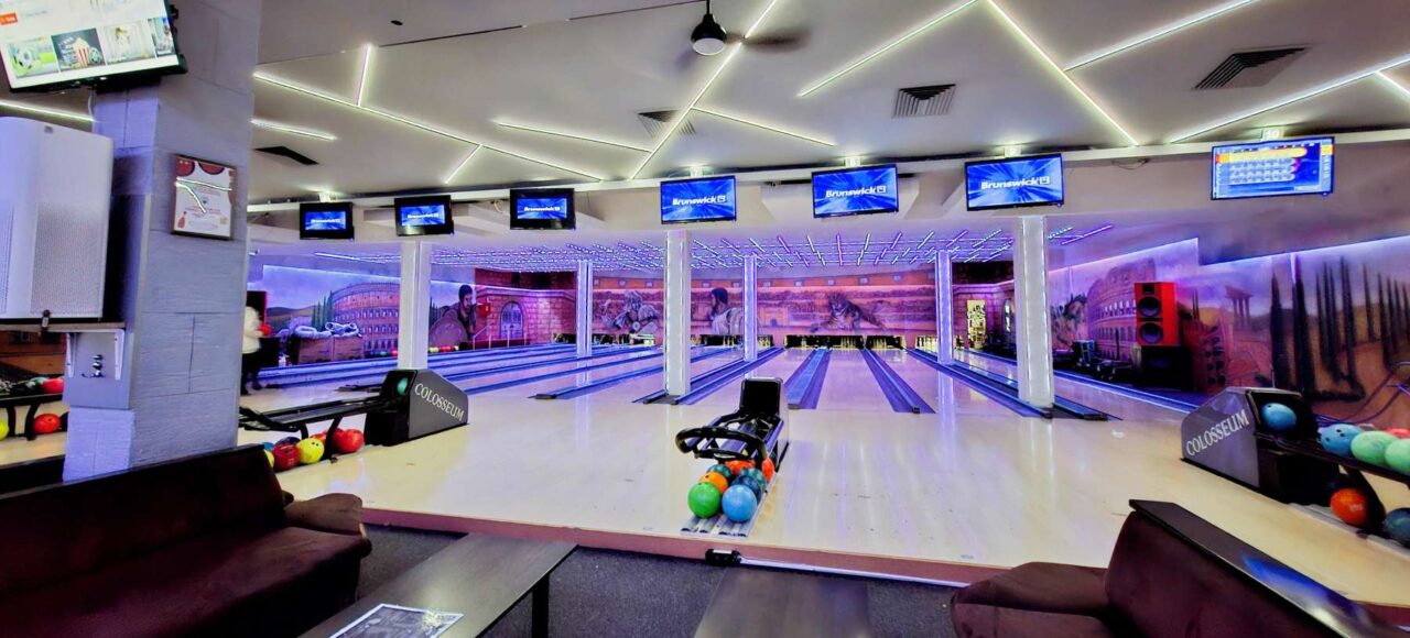 Panoramski pogled na enterijer Colosseum Bowling centra, obuhvata više staza za kuglanje, stolove za sedenje i monitori za rezultate.