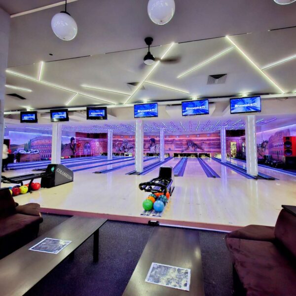 Panoramski pogled na enterijer Colosseum Bowling centra, obuhvata više staza za kuglanje, stolove za sedenje i monitori za rezultate.