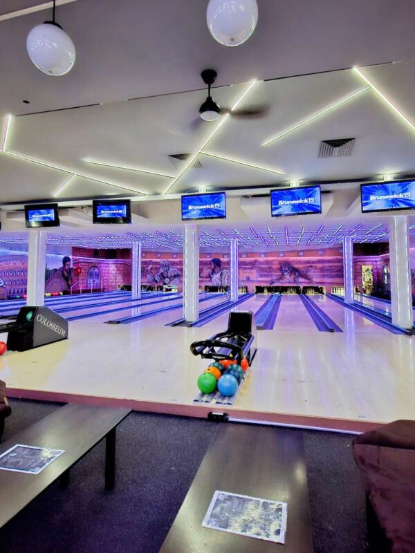 Panoramski pogled na enterijer Colosseum Bowling centra, obuhvata više staza za kuglanje, stolove za sedenje i monitori za rezultate.