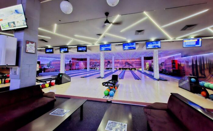 Panoramski pogled na enterijer Colosseum Bowling centra, obuhvata više staza za kuglanje, stolove za sedenje i monitori za rezultate.