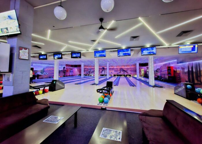 Panoramski pogled na enterijer Colosseum Bowling centra, obuhvata više staza za kuglanje, stolove za sedenje i monitori za rezultate.