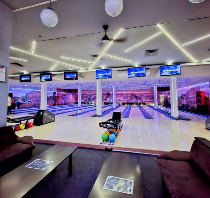 Panoramski pogled na enterijer Colosseum Bowling centra, obuhvata više staza za kuglanje, stolove za sedenje i monitori za rezultate.