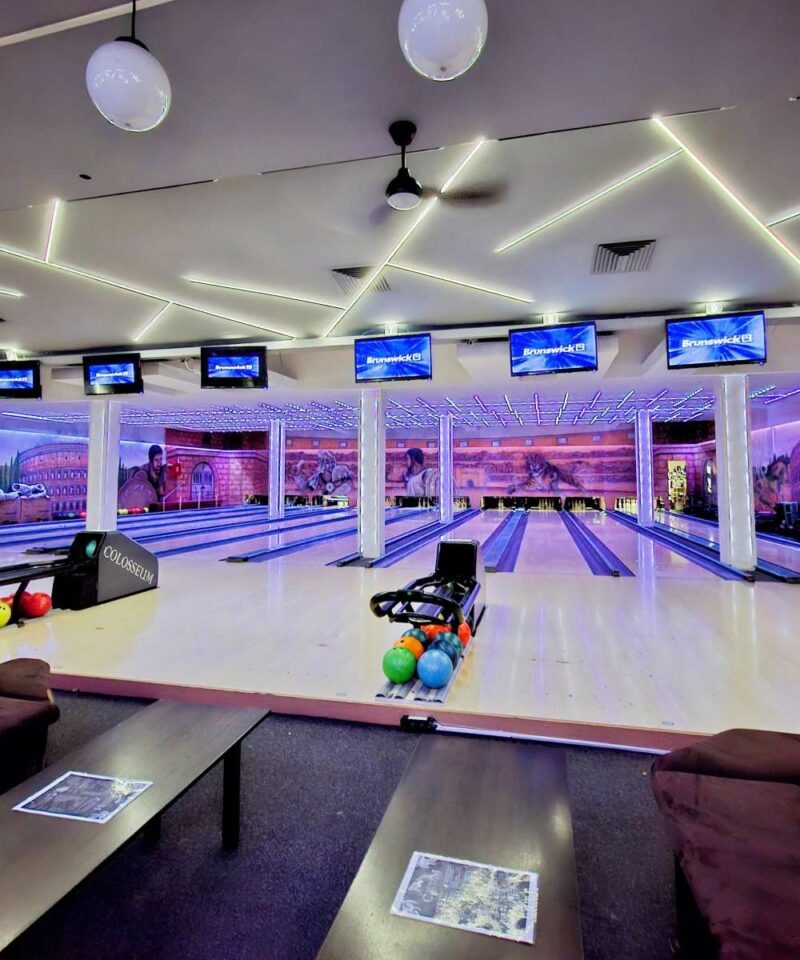 Panoramski pogled na enterijer Colosseum Bowling centra, obuhvata više staza za kuglanje, stolove za sedenje i monitori za rezultate.