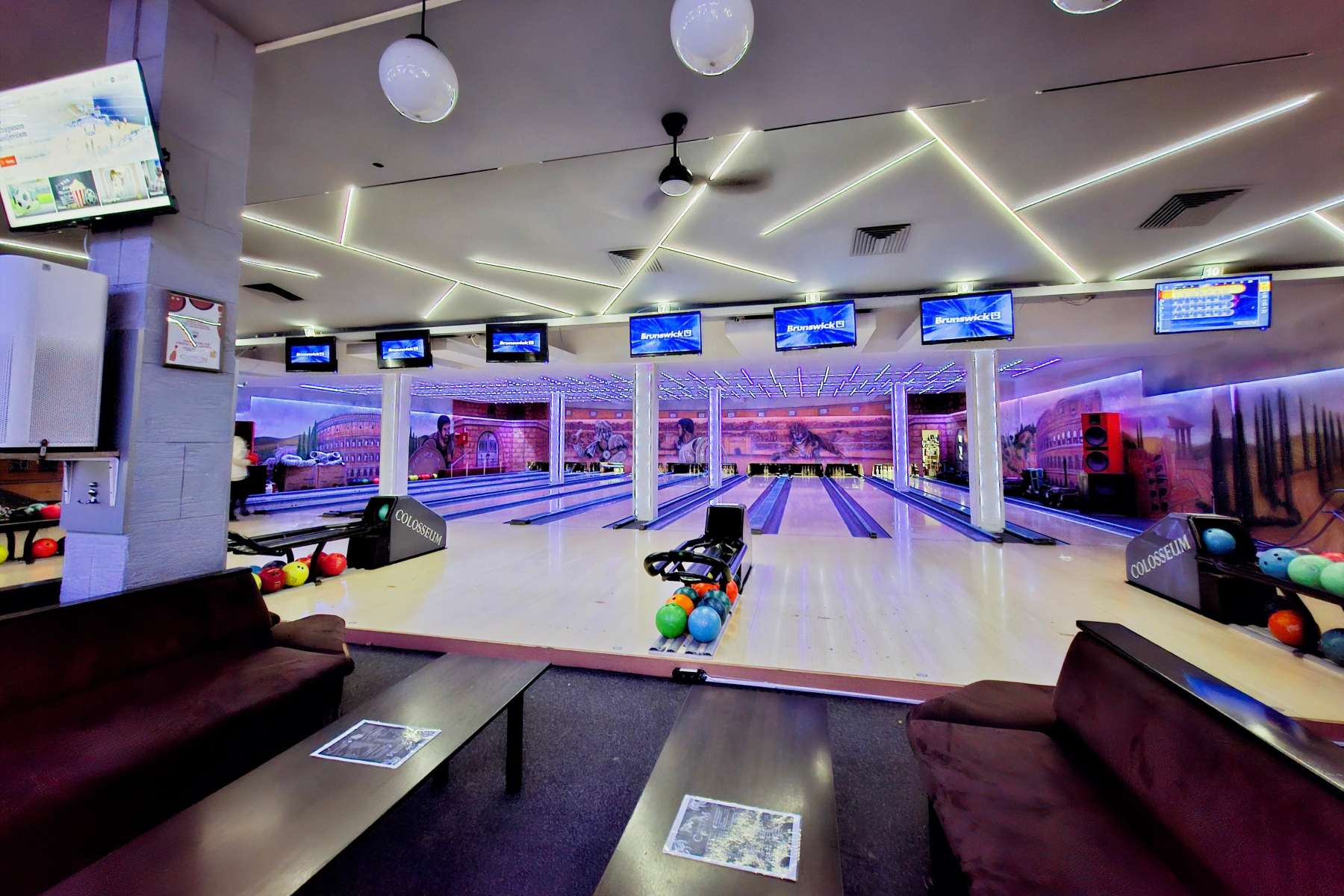Panoramski pogled na enterijer Colosseum Bowling centra, obuhvata više staza za kuglanje, stolove za sedenje i monitori za rezultate.