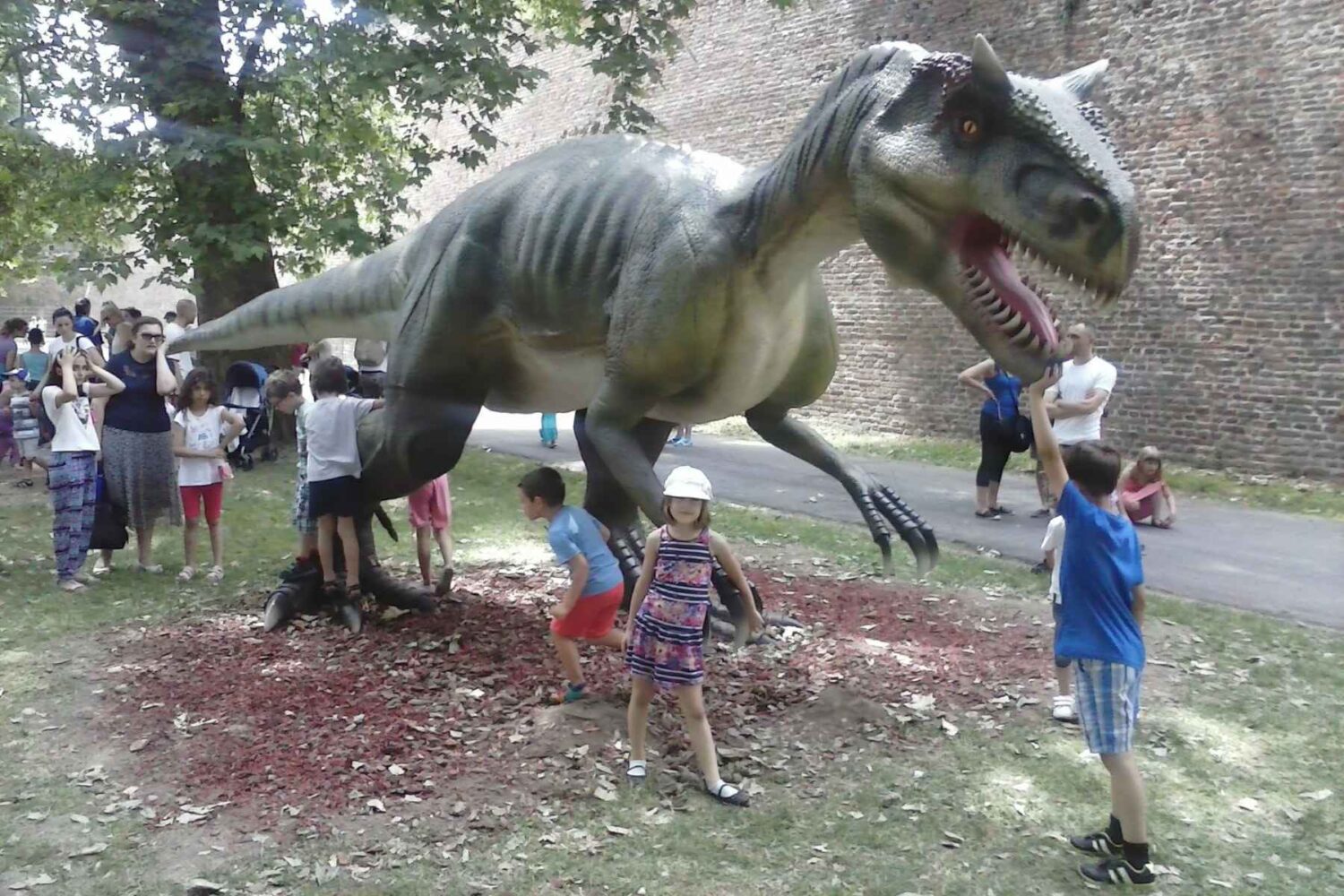 Velika figura (eksponat) dinosaura, sivkaste ili braon boje, sa zastrašujućim zubima, u prirodnom okruženju Dino parka.