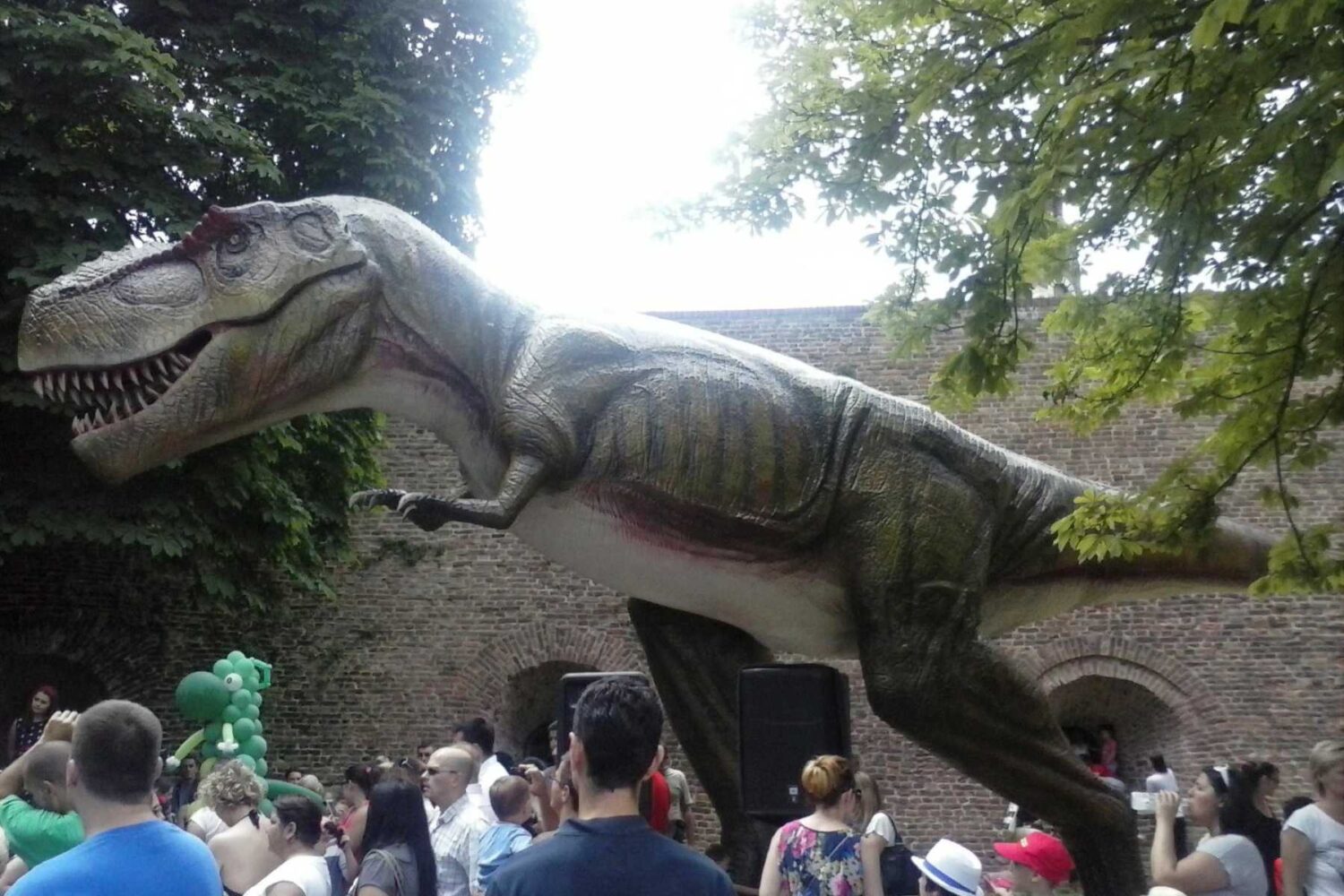 Figura dinosaura sa dugim vratom (ili oklopom), zelene ili žute boje, koja se nadvija nad drvećem/lišćem u Dino parku "Jura avantura".
