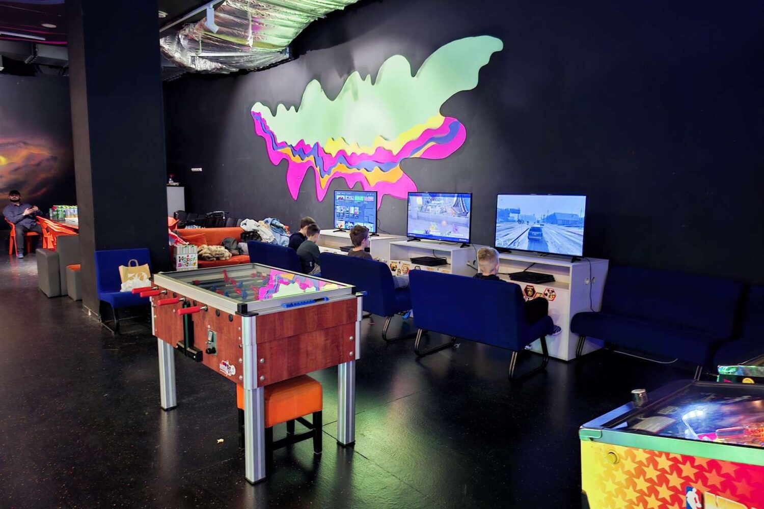 Deca (ili mlađi tinejdžeri) sede i igraju video igre na velikoj konzoli (arkadnoj mašini) sa džojsticima, u Futurama Gejming Centru.