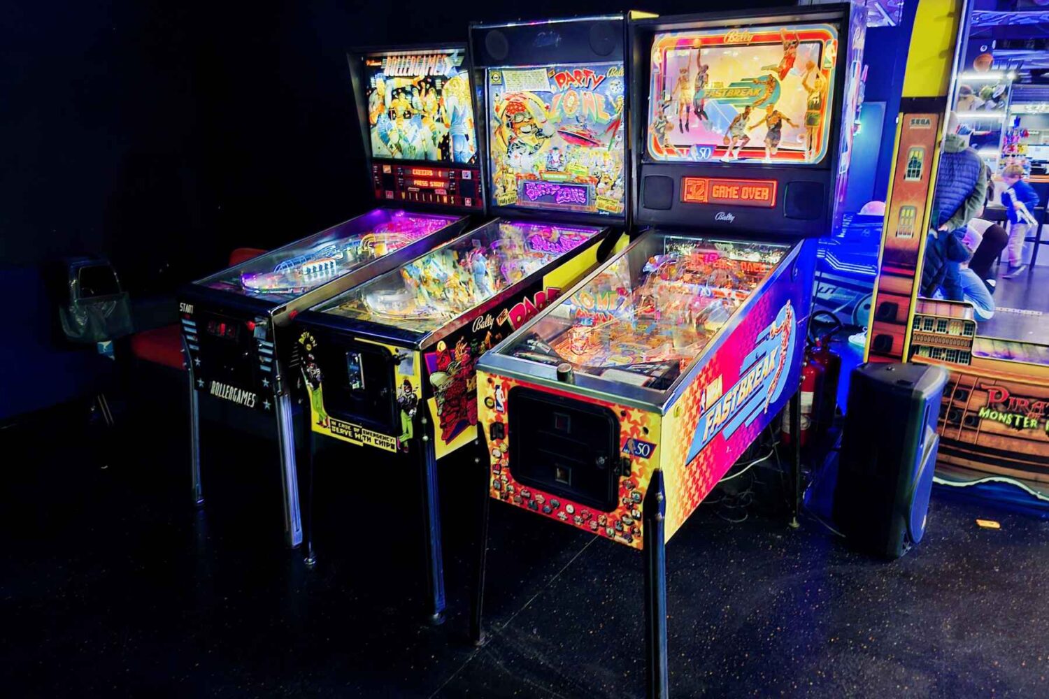 Detaljan prikaz tri fliper mašine (Pinball) – NBA Fastbreak, The Party Zone i Rollergames – sa svetlećim tablama i aktivnom lopticom.