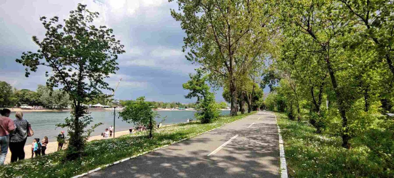 Biciklistička staza na Adi Ciganliji, okružena zelenilom, pored jezera sa bistrom vodom.