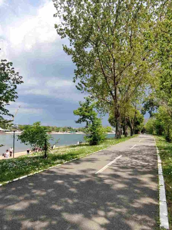 Biciklistička staza na Adi Ciganliji, okružena zelenilom, pored jezera sa bistrom vodom.