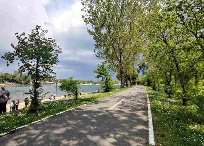 Biciklistička staza na Adi Ciganliji, okružena zelenilom, pored jezera sa bistrom vodom.