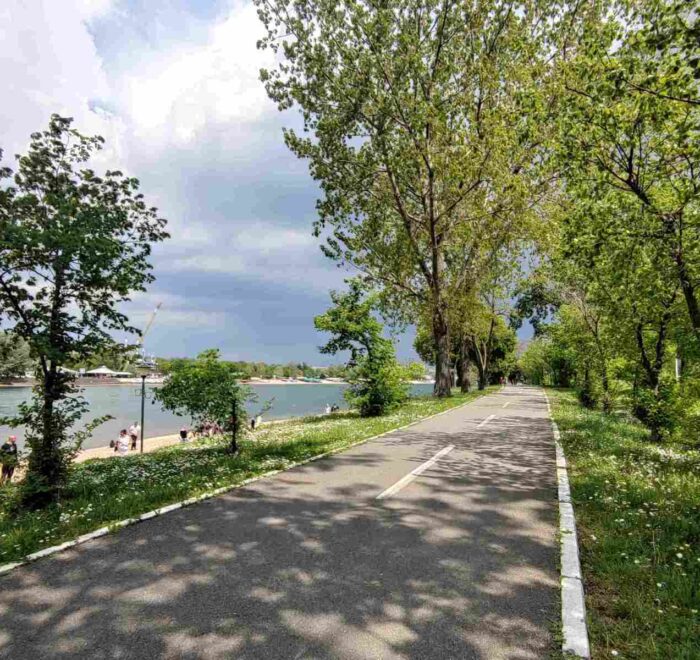 Biciklistička staza na Adi Ciganliji, okružena zelenilom, pored jezera sa bistrom vodom.