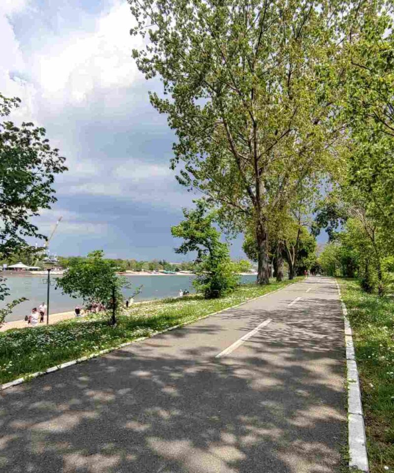 Biciklistička staza na Adi Ciganliji, okružena zelenilom, pored jezera sa bistrom vodom.