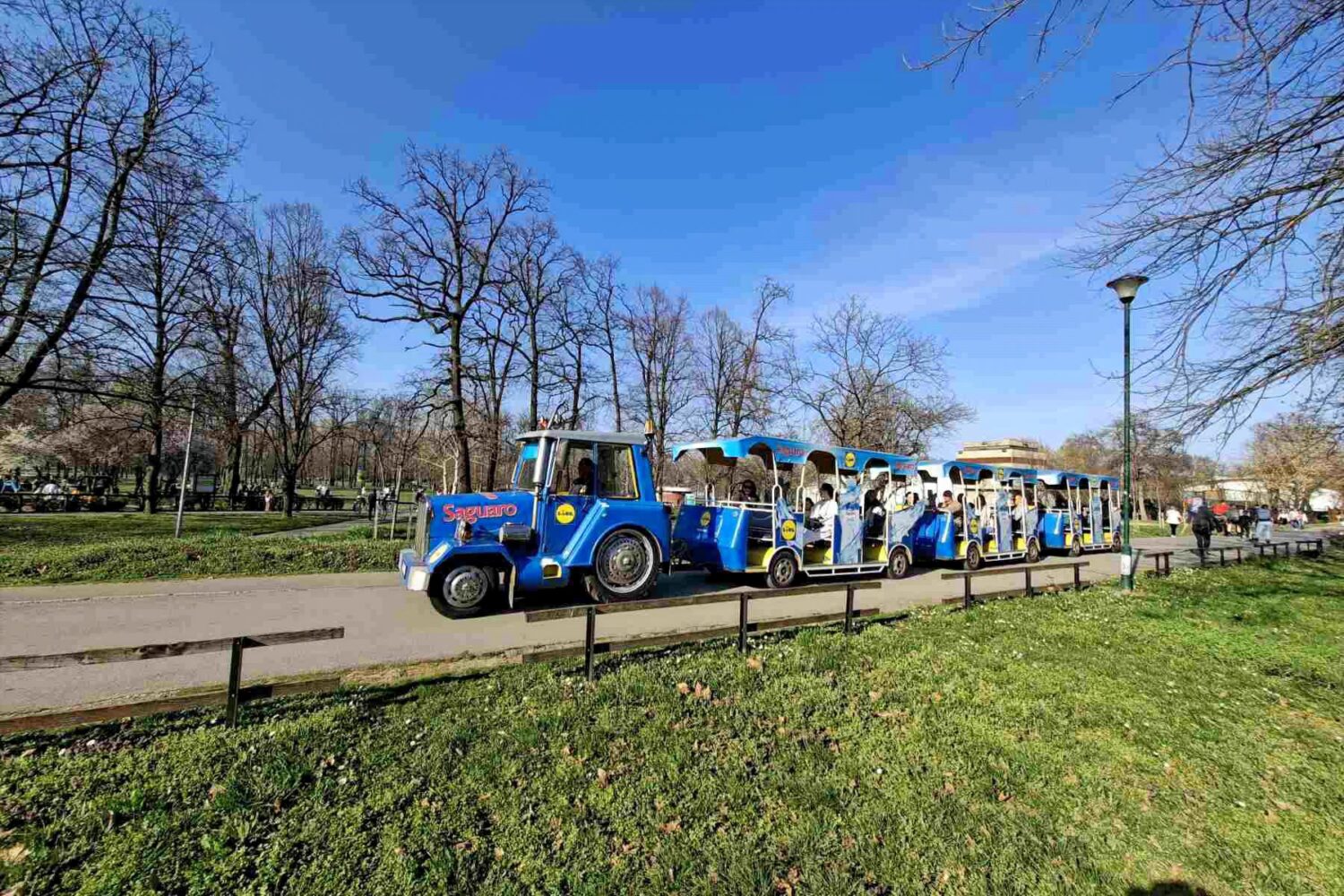 Turistički vozić na Adi Ciganliji na stazi kod novobeogradske strane okružen zelenilom i šetačima