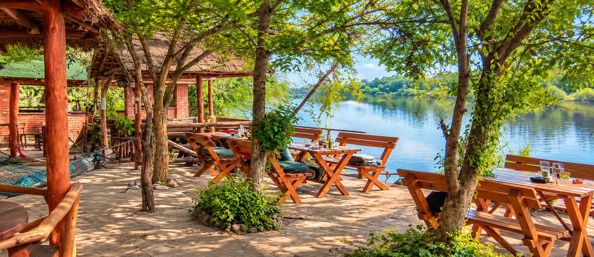 Restoran na obali reke okružen drvećem, sa drvenim stolovima i pogledom na vodu